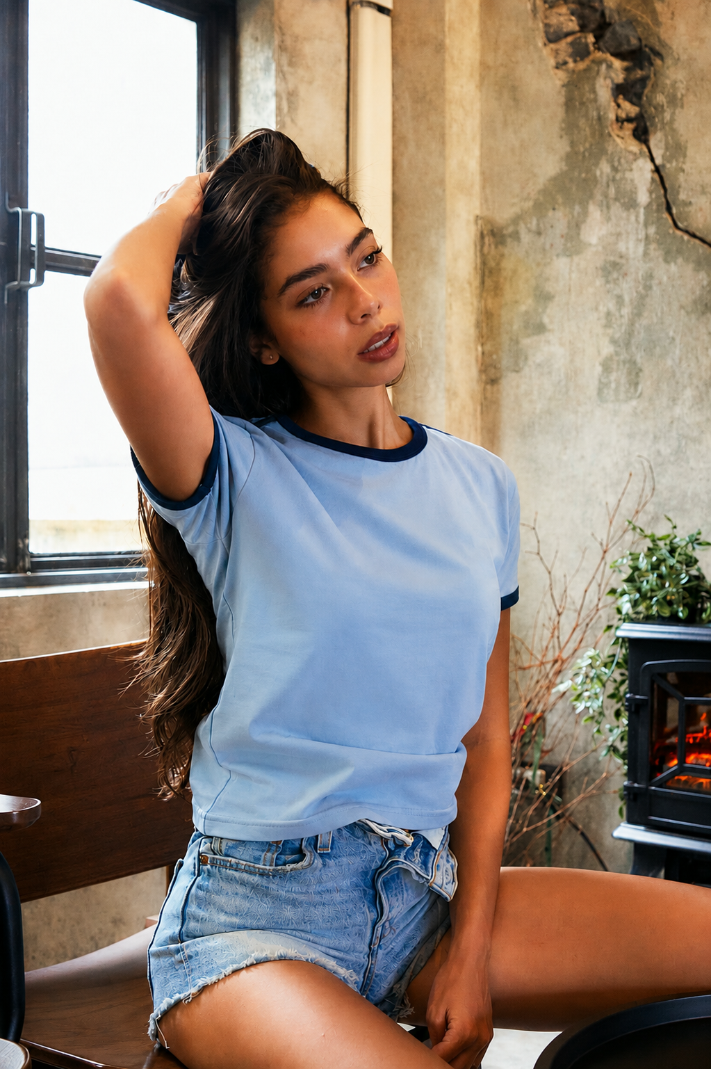 A woman wearing a light blue ringer t-shirt with dark navy trim and denim shorts, posing in a rustic room with a concrete wall and a window.