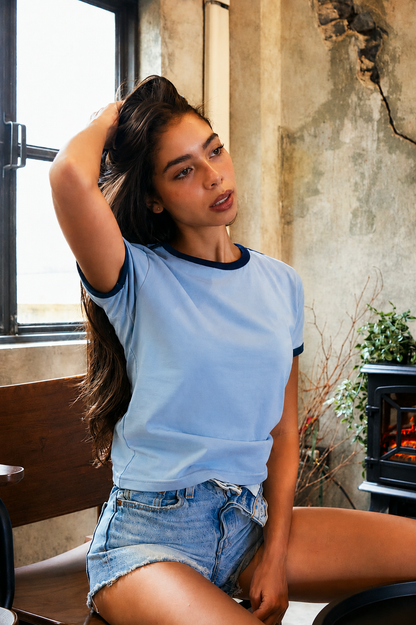 A woman wearing a light blue ringer t-shirt with dark navy trim and denim shorts, posing in a rustic room with a concrete wall and a window.