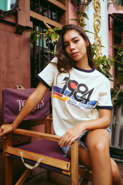 A woman sits on a wooden chair with a purple cushion, wearing a white ringer T-shirt with navy trim and a large multicolor graphic on the front that reads 'TGA Los Angeles'.