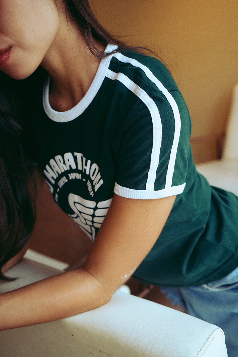 A close-up lifestyle shot of a woman wearing a dark green ringer t-shirt with white stripes on the sleeves and a white graphic print on the front that reads 'MARATHON'.