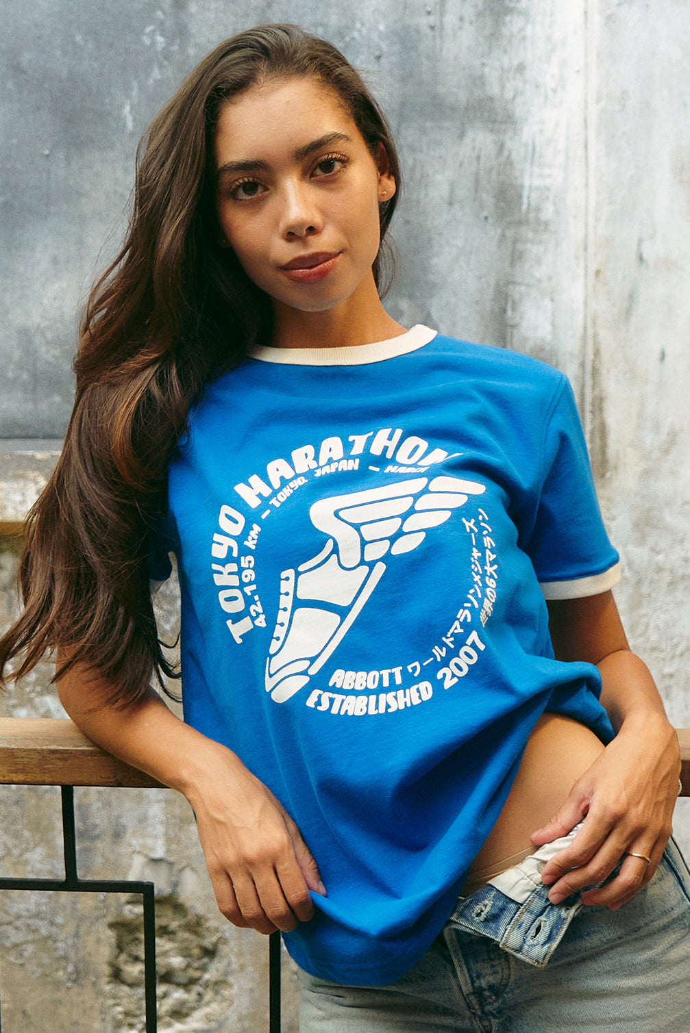 A woman wearing a bright blue short-sleeve graphic T-shirt with white trim and a large white chest print reading 'Tokyo Marathon' and 'Abbott Established 2007', posed against a textured gray wall.