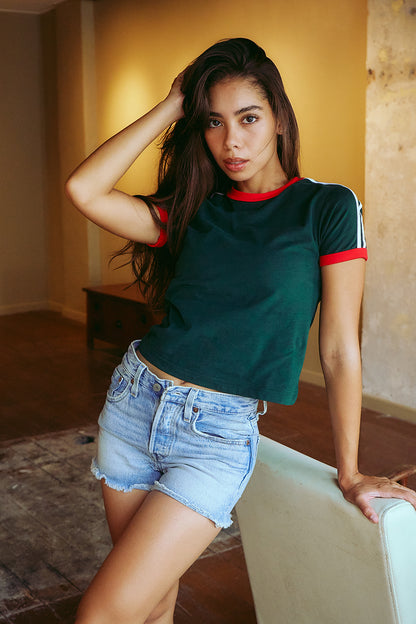 A woman posing indoors wearing a dark green ringer t-shirt with red and white trim on the collar and sleeve cuffs, paired with light blue denim shorts.