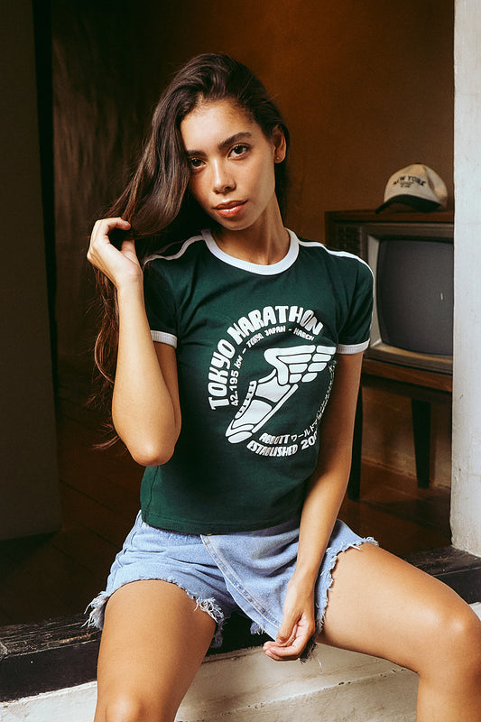 A woman wearing a dark green Tokyo Marathon ringer t-shirt with white text and a sneaker graphic, paired with light blue denim shorts, sitting in a dimly lit room.