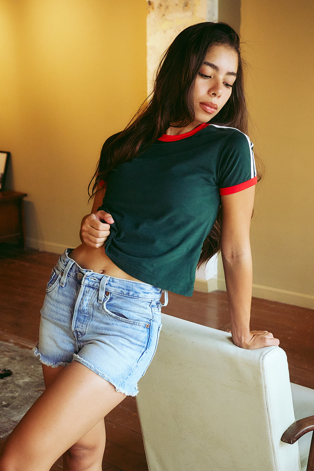 A woman posing indoors wearing a dark green ringer t-shirt with red and white trim, paired with light-wash denim shorts, leaning against a white chair.