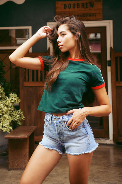 A woman posing in a dark green ringer t-shirt with red trim and light wash denim shorts in an outdoor cafe setting.