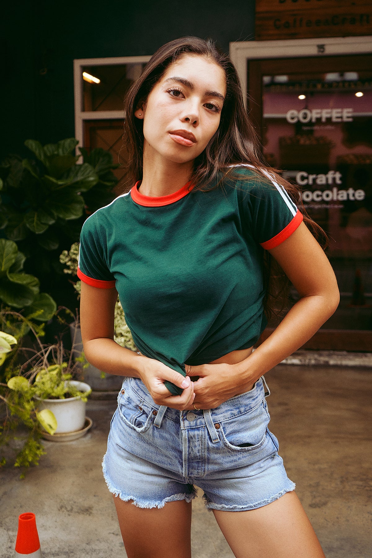A woman wearing a dark green ringer t-shirt with red trim and light blue denim shorts, standing in front of a coffee shop storefront.