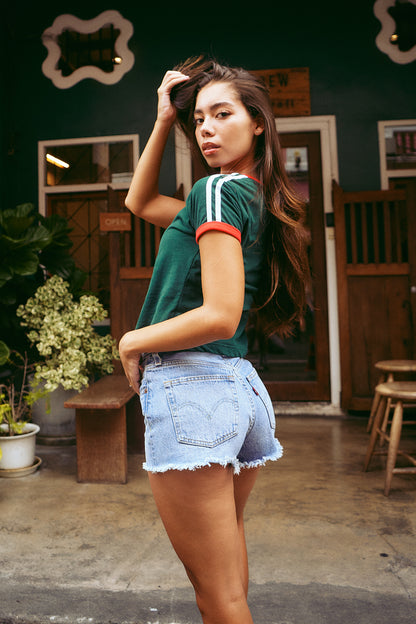 A woman posing in a dark green short-sleeve athletic t-shirt with white and red stripes on the sleeves, paired with light-wash denim shorts, set against a rustic cafe background.