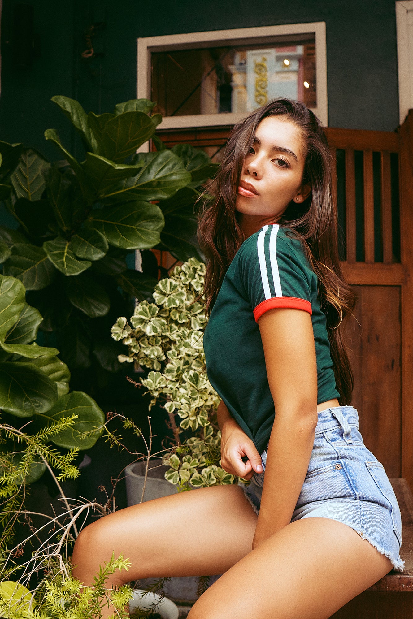 A woman sitting outdoors in front of a dark green wall and wooden door, wearing a dark green short-sleeve athletic crop top with white and red stripes on the sleeves, paired with light blue denim shorts.
