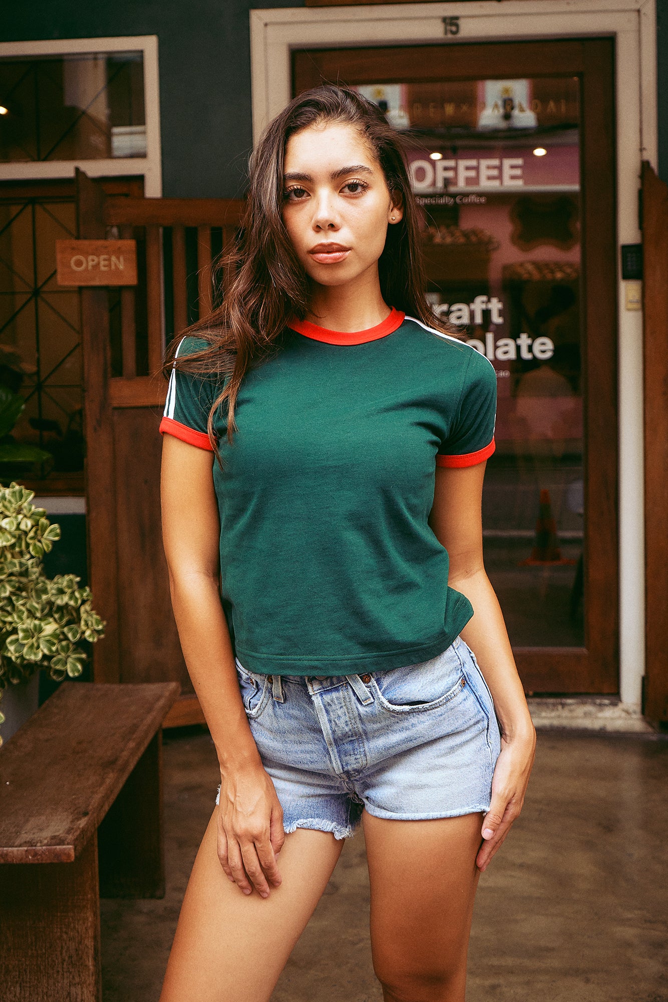 A woman modeling a dark green ringer t-shirt with red and white trim on the crew neck and sleeves, paired with light-wash denim shorts, standing in front of a rustic wooden door and shop window.