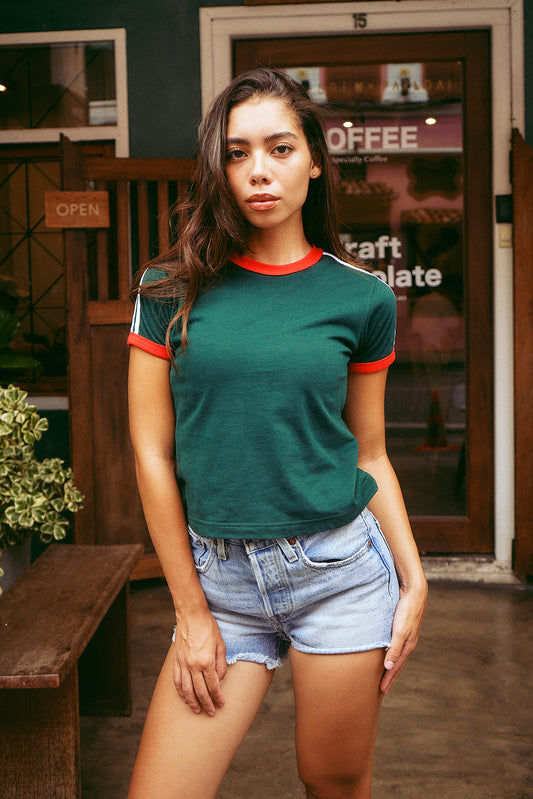 A woman modeling a dark green ringer t-shirt with red and white trim on the crew neck and sleeves, paired with light-wash denim shorts, standing in front of a rustic wooden door and shop window.