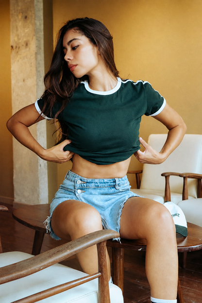 A woman sitting on a wooden chair wearing a dark green ringer t-shirt with white trim and light-wash denim shorts in a warm indoor setting.