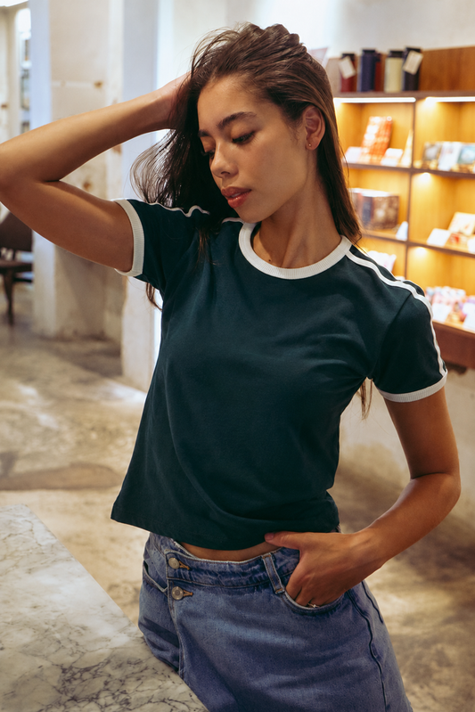 A woman wearing a dark teal ringer t-shirt with white trim on the collar and sleeve cuffs, paired with blue jeans, posing in a warmly lit indoor setting.