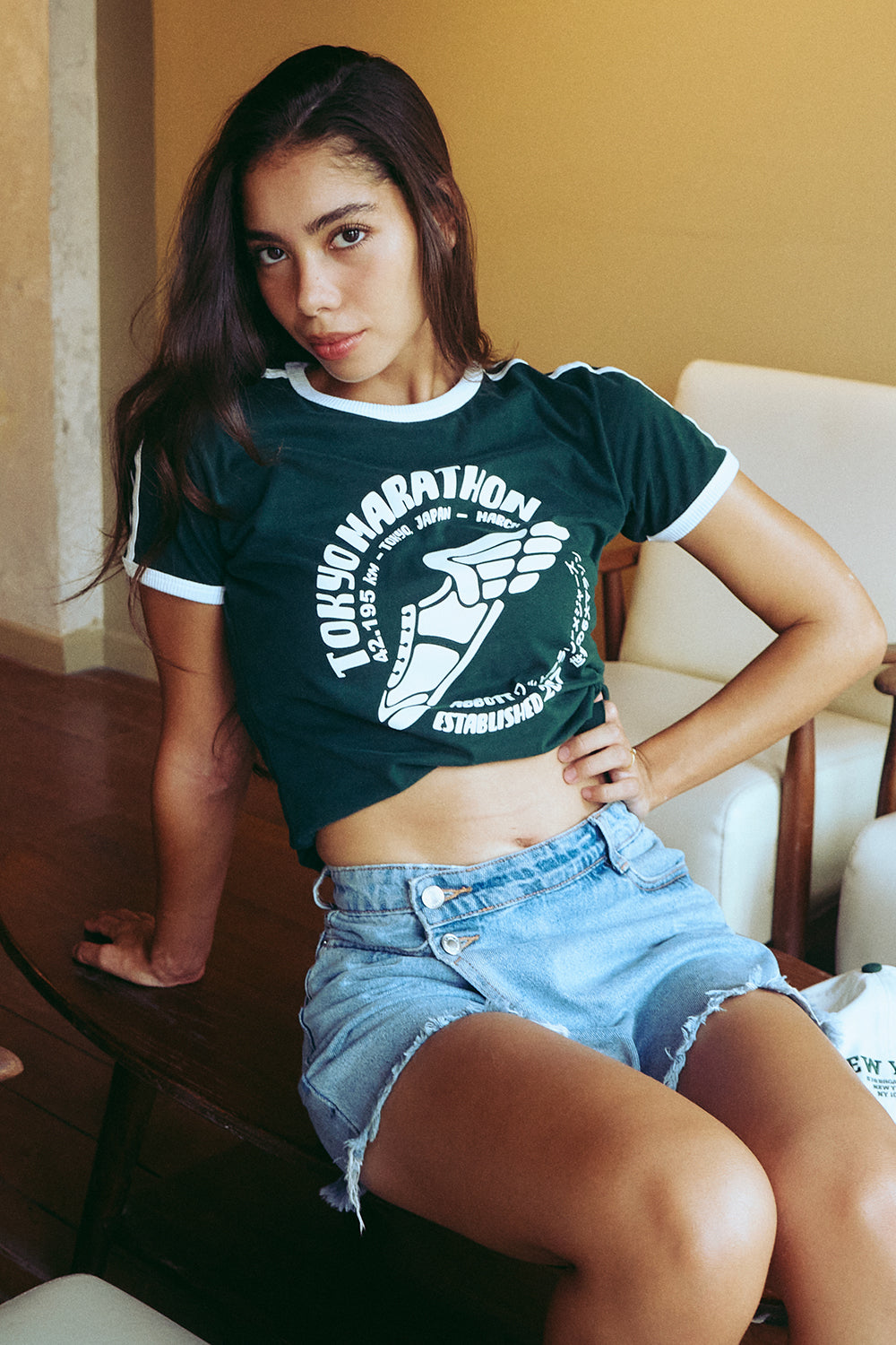 A woman wearing a dark green cropped ringer T-shirt with white trim and a white graphic print that reads 'Tokyo Marathon' and 'Est. 2016', paired with light blue denim shorts, sitting in an indoor setting.