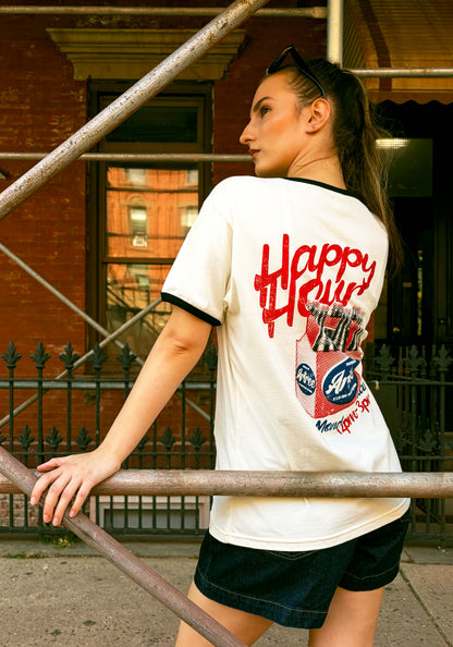 A woman wearing a white ringer t-shirt with black trim and a red 'Happy Hour' graphic on the back, leaning against a metal railing in front of a brick building.