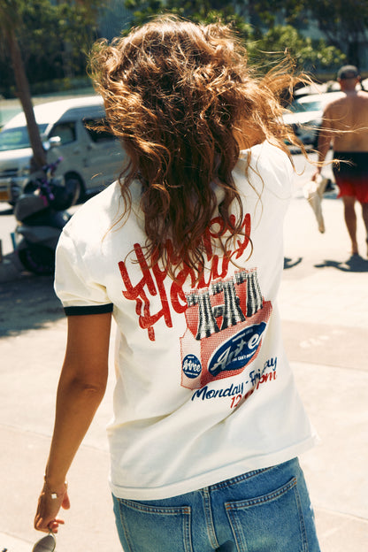 A person viewed from behind wearing a white ringer T-shirt with black trim and a large red and blue graphic print on the back, paired with blue jeans in an outdoor street setting.