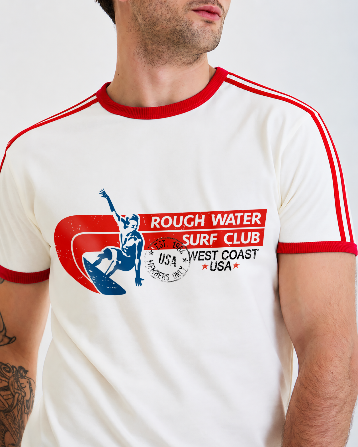 A man wearing a white ringer t-shirt with red trim and a red, blue, and black graphic on the chest that reads 'ROUGH WATER SURF CLUB' and 'WEST COAST USA'.